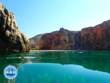 Blick vom kristallklaren türkisfarbenen Wasser auf den Strand der Agiofarango Schlucht an der Südküste von Kreta, Griechenland, umgeben von hohen Felswänden unter blauem Himmel.