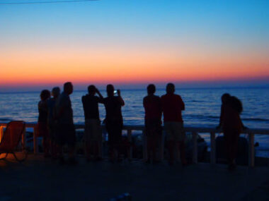 Gruppe von Urlaubern beobachtet einen farbenfrohen Sonnenuntergang über dem Meer von der Terrasse der Zorbas Island Apartments auf Kreta, Griechenland.