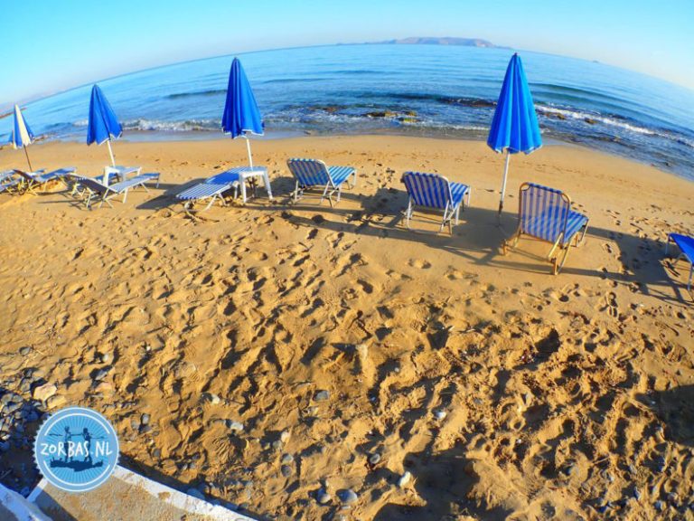 Sandstrand von Kokkini Hani auf Kreta mit blauen Sonnenschirmen und Blick auf das Mittelmeer bei Zorbas Island.
