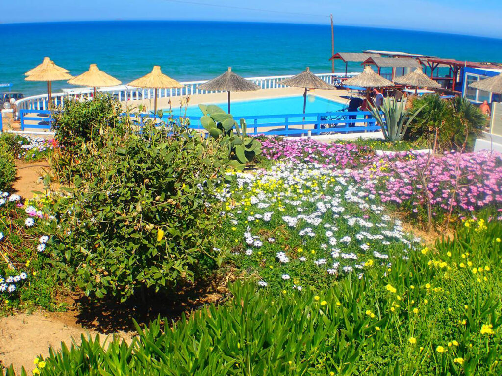 Bunter Blumengarten mit rosa, weißen und gelben Blüten im Vordergrund, dahinter ein Swimmingpool mit blauem Zaun und Blick auf das Meer in Kokkini Hani.