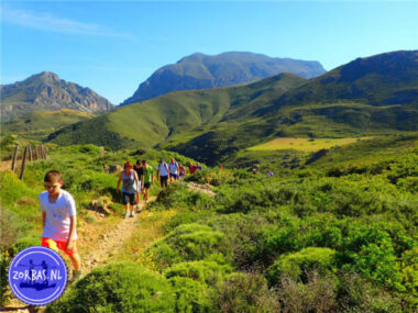 Geführte Wandergruppe auf einem Pfad durch grüne mediterrane Vegetation und Büsche vor einer beeindruckenden Bergkette unter blauem Himmel auf Kreta.