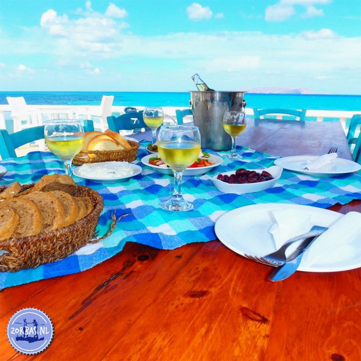 Gedeckter Holztisch mit blau-weiß karierter Tischdecke, Weißwein, frischem Brot und Oliven vor der Kulisse des türkisblauen Meeres in Kokkini Hani, Kreta.