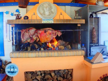 Traditionelles griechisches Barbecue mit einem ganzen Lamm am Drehspieß über offenem Holzfeuer in der Außenküche von Zorbas Island, Kreta, unter einem Kalimera-Schild.