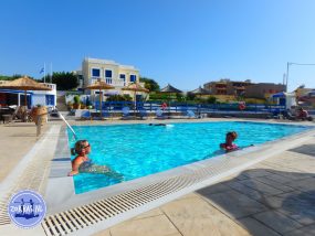 Urlauber entspannen in einem großen Außenpool einer Hotelanlage auf Kreta unter blauem Himmel mit Blick auf traditionelle Gebäude.